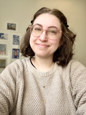 Image of Shiloh Soto, a woman with shoulder-length brown hair, beige translucent transitions lenses, a comfy beige sweater, and gold hoop earrings and a simple gold necklace. She's smiling and content
